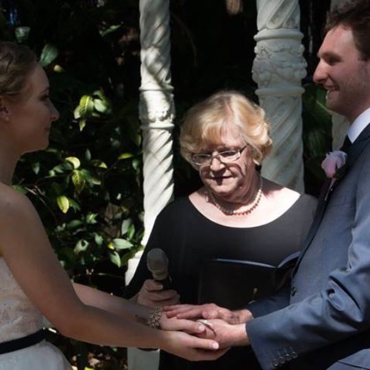Jennifer Cram, Inclusive Marriage Celebrant
                      conducting a marriage ceremony. She is wearing an
                      understated black top and holds an unadorned A5
                      black folder and a black microphone. The bride and
                      groom are holding hands.