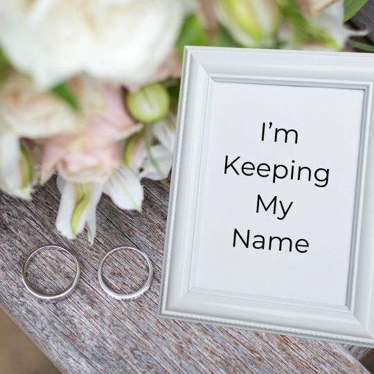 Silver wedding rings and a silver framed
                      graphic that reads "I'm Keeping My Name"
                      on weathered gray boards with pastel wedding
                      flowers in the background