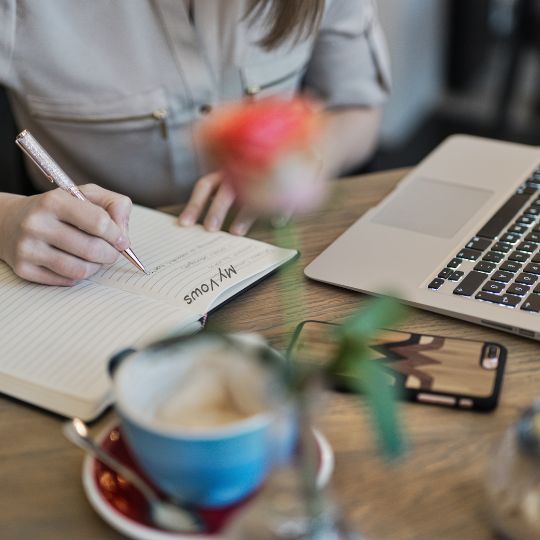 Woman using
                      a yellow pencil to writing vows in a notebook . A
                      blue cup of coffee on a red saucer, a laptop, a
                      mobile phone, and an out of focus pink rose in a
                      bud vase are also on the table