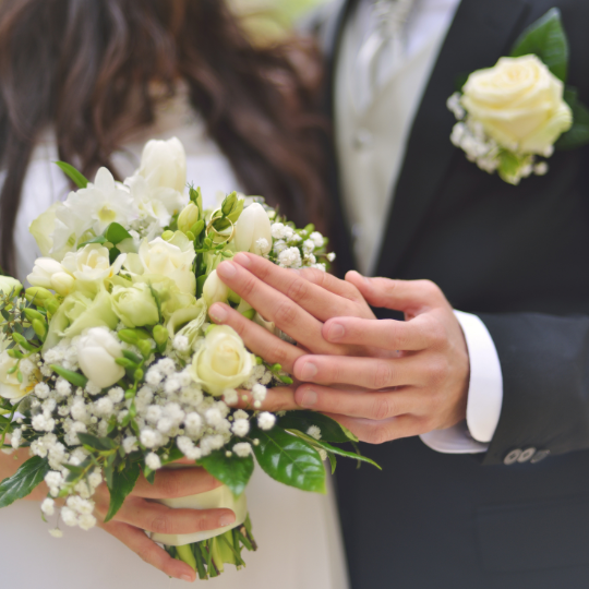 Close up of
                      bride and groom's hands. Groom holding the bride's
                      hand, bride holding a bouquet of white roses and
                      lily of the valley. The groom wears a dark suit, a
                      silver gray tie and has a white rose boutonniere
                      on his left lapel