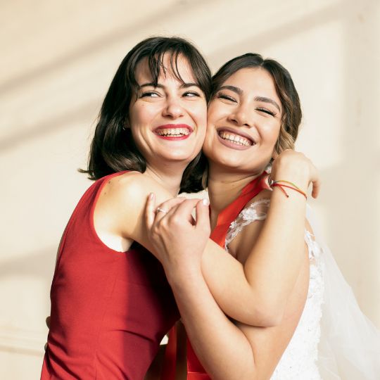 Bride hugging BFF.
                      Bride is wearing white. BFF is wearing a red dress
                      with spaghetti straps