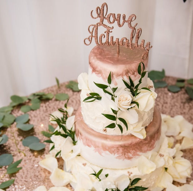 Wedding Cake with Love Actually topper
                      standing on a table covered in white rose petals
                      and green leaves