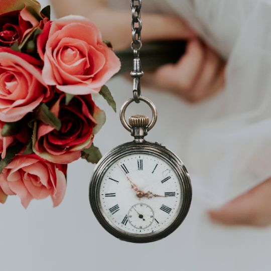 A silver pocket
                        watch showing the time in Roman Numerals as 5
                        minutes to 3 with a bride holding a bouquet of
                        dark pink roses in the background